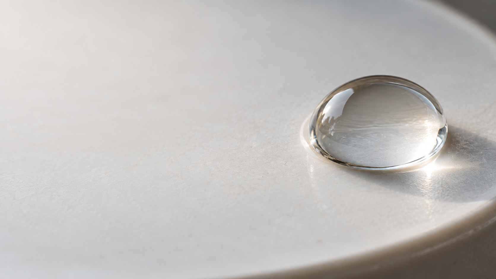 A single large clear water droplet sits on matte pale porcelain near the right edge, minute meniscus and accurate refraction.