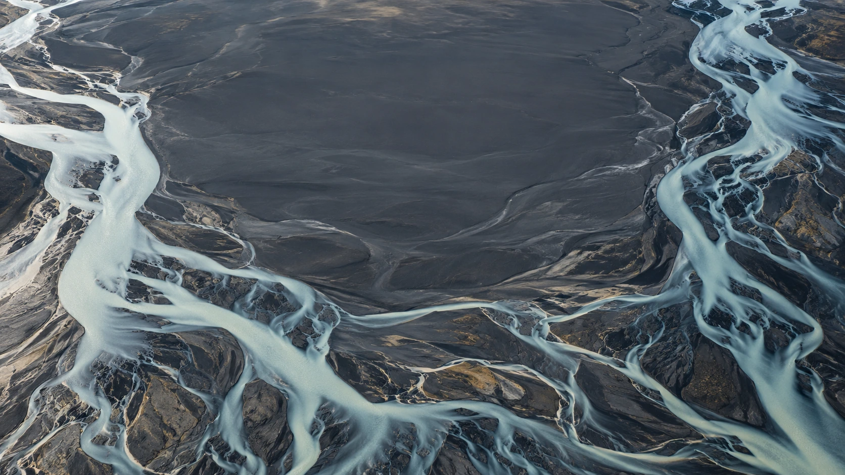 Overhead view of pale milky glacial rivers braiding across dark volcanic sand, fine branching details near the lower half.